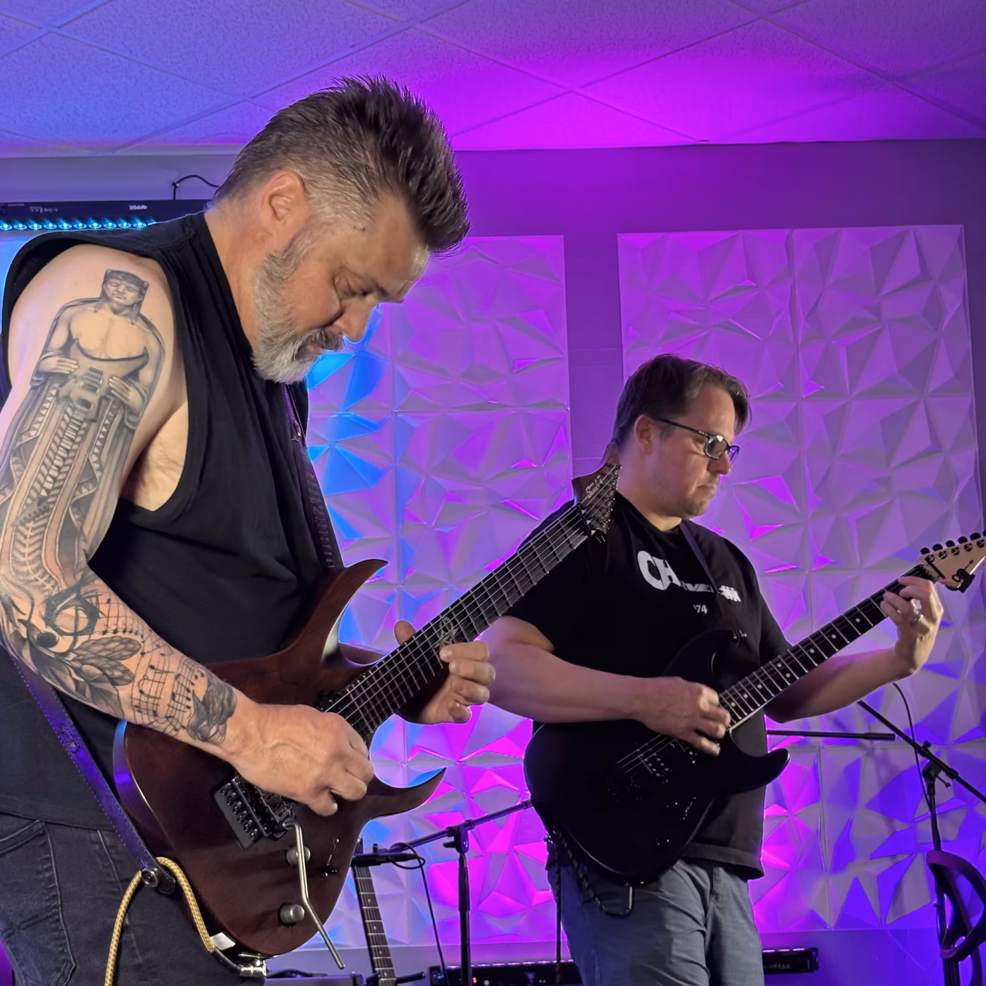 Two guitarists playing electric guitars during a Silversound Guitar open jam session, with purple and blue stage lighting and acoustic panel walls in the background.